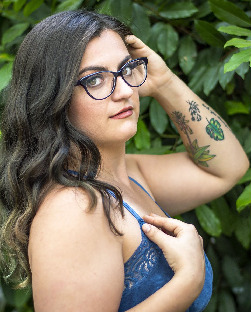 Maddie against a wall of green leaves, glasses on, one hand in her hair, looking back over her shoulder